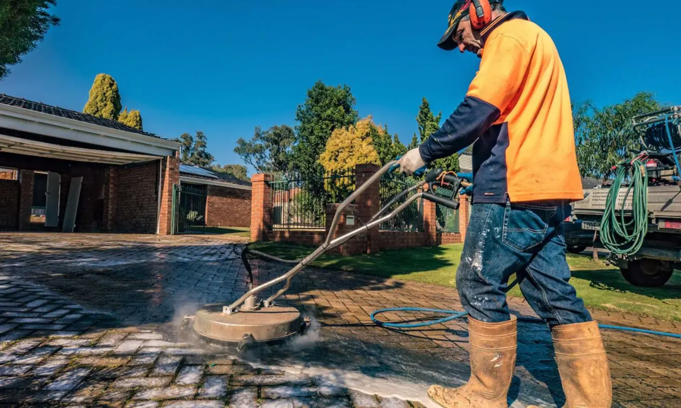 A man in an orange shirt operates a pressure washer to clean a brick driveway, removing dirt and stains effectively.